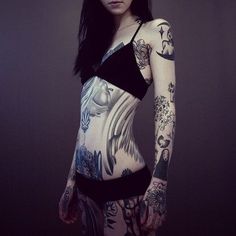 a woman with tattoos on her body standing in front of a dark background and looking at the camera