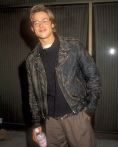 a man in glasses and a leather jacket is holding a bottle of vodka while standing next to a wall