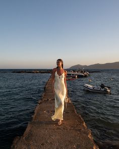 a woman in a white dress is walking along the edge of a pier near boats