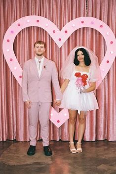 a man and woman standing in front of a heart shaped photo booth with pink curtains