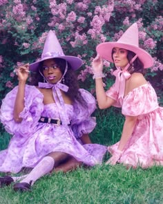 two women in dresses and hats sitting on the grass with purple flowers behind their backs