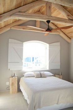 a white bed sitting under a window in a bedroom next to a wooden ceiling fan