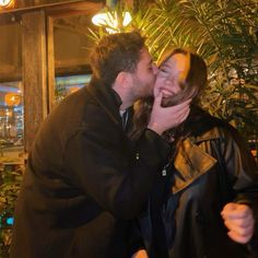 a man and woman kissing each other in front of a potted plant at night