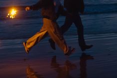 two people running on the beach at night with their feet in the air and one person holding an umbrella