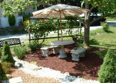 there is a table and two benches under an umbrella in the yard with gravel on the ground