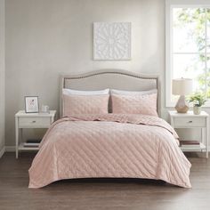 a bed with pink comforter and pillows in a white room next to a window