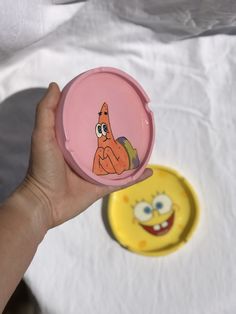 a hand holding a pink spongebob compact container next to a yellow spongebob plate