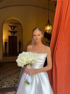 a woman in a white dress holding a bouquet of flowers next to an orange curtain