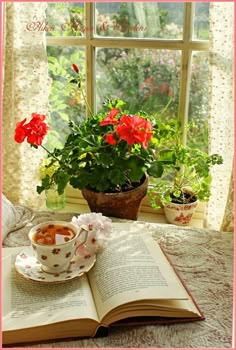 an open book and cup on a table next to a window with flowers in it