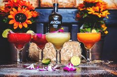 three different colored cocktails sitting on top of a table next to bottles and flowers