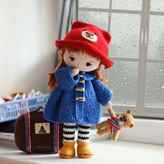 a knitted doll is holding a small dog and wearing a red hat, blue coat and striped pants