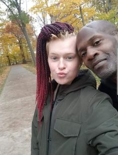 a man and woman standing next to each other in front of trees with leaves on the ground