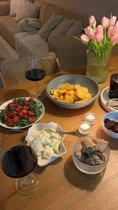 a wooden table topped with plates and bowls of food next to glasses of red wine