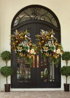 two decorative wreaths on the front door of a house with potted plants and trees