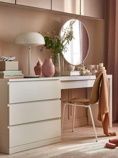 a white dresser topped with pink vases next to a mirror and table lamp on top of it