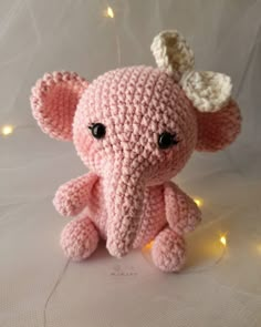 a small pink crocheted elephant sitting on top of a white sheet with lights around it