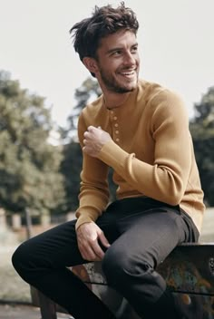 a young man sitting on top of a bench in a park wearing a yellow sweater and black pants