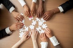 several people are holding hands together to make a puzzle on a wooden table with white jigsaw pieces