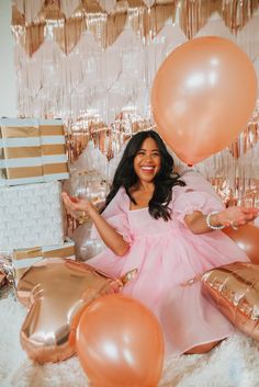 a woman in a pink dress sitting on a bed with balloons and presents around her