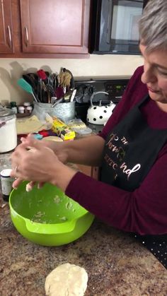 a woman in an apron mixing dough into a green bowl on top of a counter