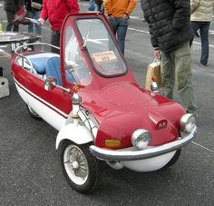 a small red and white car parked on the side of a road next to people