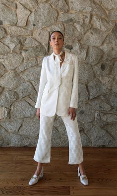 a woman standing in front of a stone wall wearing white pants and a blazer