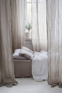 an unmade bed with white sheets and pillows in front of a window covered by sheer curtains