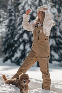 a woman standing in the snow next to a brown dog wearing overalls and a hat