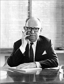 an older man sitting at a desk talking on the phone and holding his hand to his face