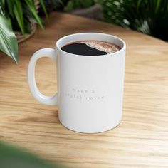 a white coffee mug sitting on top of a wooden table next to a green plant