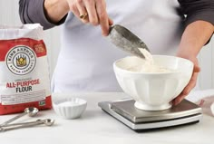 a person in an apron is mixing flour into a white bowl with a measuring spoon