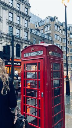 a woman standing next to a red phone booth on the sidewalk in front of tall buildings