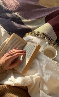 a person is reading a book and drinking coffee on a bed with blankets around them