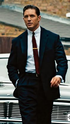 a man in a suit and tie walking next to a car with his hands in his pockets