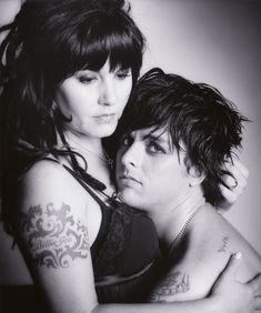 black and white photograph of two women hugging each other with tattoos on their arms, both looking at the camera