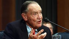 a man in a suit and tie sitting at a table with a microphone behind him