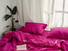 an unmade bed with bright pink sheets and pillows in front of a large window