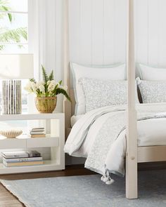 a bed with white sheets and pillows next to a plant in a vase on a table