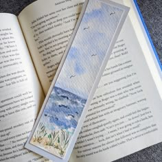 an open book with a watercolor painting on it next to an open bookmark