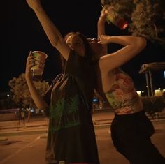 two people standing in the street holding up cups and drinking beverages at night time with palm trees behind them