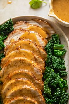 a white plate topped with meat and broccoli covered in gravy next to a bowl of sauce