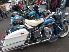 several motorcycles are parked on the side of the road in a parking lot with people standing around