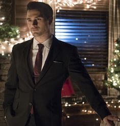 a man in a suit and tie standing next to a christmas tree with lights on it