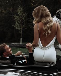 a woman in a white dress is sitting in a black convertible car with a man standing next to her