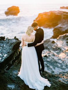 a bride and groom standing on the rocks by the ocean at sunset in their wedding attire