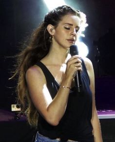 a woman holding a microphone in her right hand and looking down at the ground with lights behind her