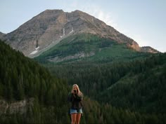 a woman standing on top of a lush green hillside next to a forest covered mountain