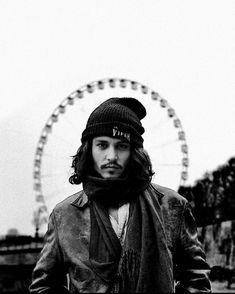 a man standing in front of a ferris wheel wearing a beanie hat and scarf