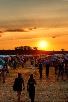 a crowd of people walking across a grass covered field next to tents and trees at sunset