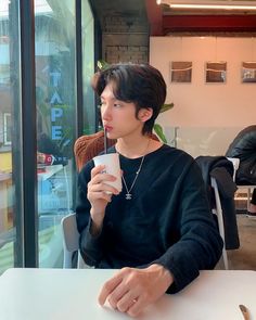 a young man sitting at a table with a drink in his hand and looking out the window